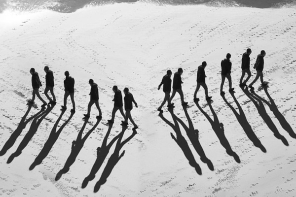 Schwarz-weiße Aufnahme von zwei Personengruppen, die auf sandigem Untergrund in getrennte Richtungen gehen, mit langen Schatten im Vordergrund