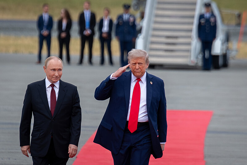 Das Foto zeigt Präsident Wladimir Putin, wie er neben Präsident Donald Trump auf einem roten Teppich auf dem Rollfeld eines Flughafens geht. Putin lächelt, während Trump etwas ernster wirkt und seinen rechten Arm in einer Art militärischem Gruß an die Stirn hält.
