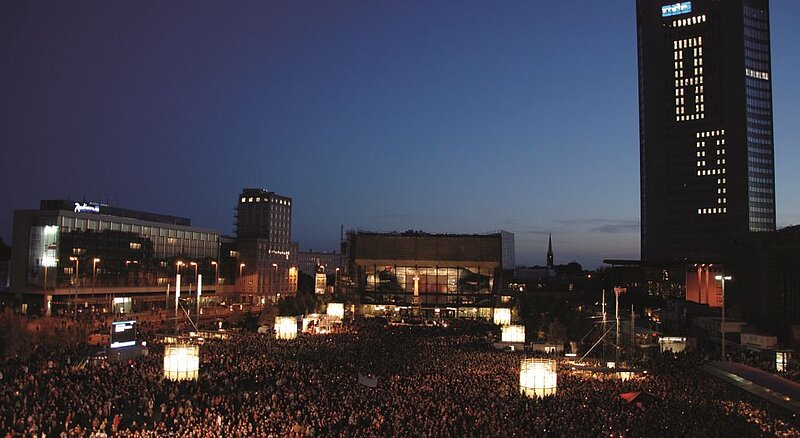 Menschenmassen auf dem Augustplatz in Leipzig am Abend: Gedenkfeier am 9. Oktober 2009 anlässlich des 20. Jahrestags der Friedlichen Revolution; im Hintergrund hohe Gebäude und der dunkle Abendhimmel zu sehen; auf dem Hochhaus rechts steht eine 89