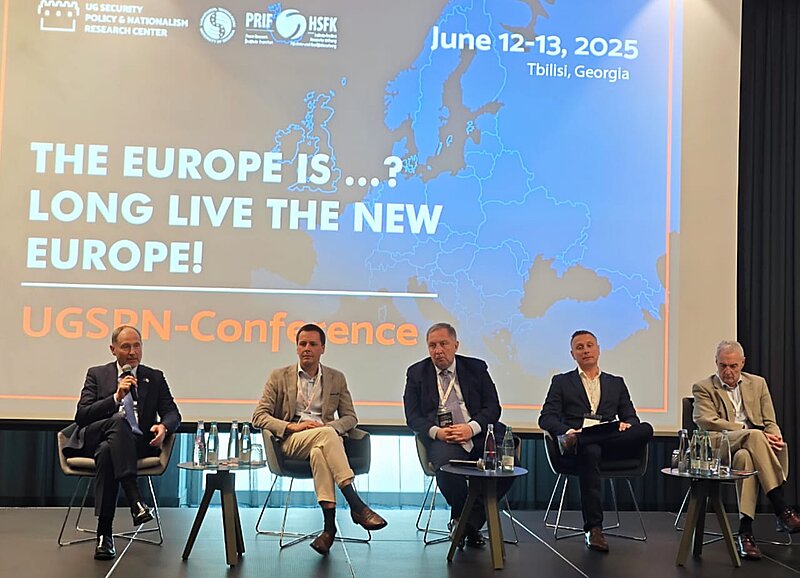 Fünf männliche Teilnehmer sitzen auf Stühlen vor einer Leinwand mit dem Konferenztitel „THE EUROPE IS …? LONG LIVE THE NEW EUROPE!