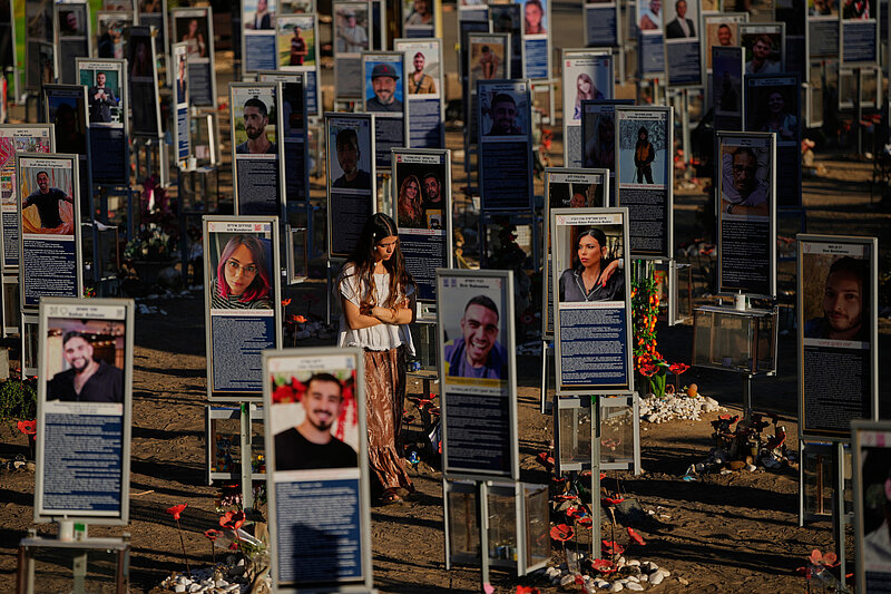Die Szene zeigt eine Gedenkveranstaltung für die Opfer des Hamas-Angriffs auf das Nova-Festival am 7. Oktober 2023. Im Mittelpunkt stehen zahlreiche Gedenktafeln mit Porträtfotos und Texten, die aufrecht in Reihen angeordnet sind. Eine Person mit langem Haar und heller Kleidung steht zwischen den Tafeln. Der Boden ist mit Blumen und Kerzen bedeckt.