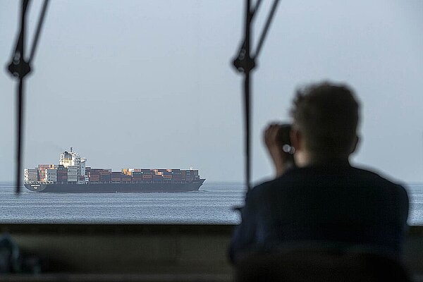 Fotografie eines Containerschiffes auf offener See. Im Vordergrund ist die unscharfe Silhouette einer Person zu sehen, die ihr Fernglas auf das Schiff gerichtet hat.