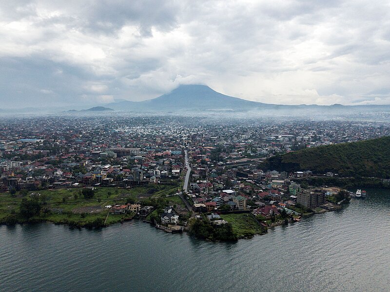 Zu sehen ist Goma, die Hauptstadt der Provinz Nord-Kivu, im Osten der Demokratischen Republik Kongo. Im Hintergrund ist ein Berg; im Vordergrund Wasser; dazwischen die Stadt