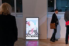 Three people are standing next to a screen. The screen shows a person washing themselves in a bathroom. The word “CLEAN” is written under the picture.