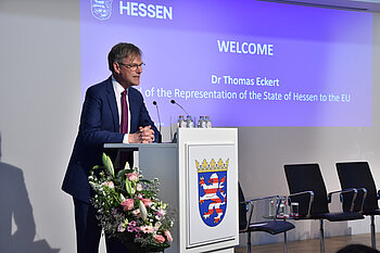 Thomas Eckert is standing at a lectern bearing the coat of arms of Hesse. The first slide of the presentation can be seen in the background.