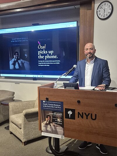 Eldad Ben Aharon is standing at a lectern. In the background, the cover of his book can be seen on a large screen.