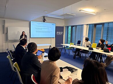 Tina Cramer spricht vor einer Gruppe. Im Hintergrund ist ein Screen und ein Aufsteller mit dem PRIF Logo zu sehen.