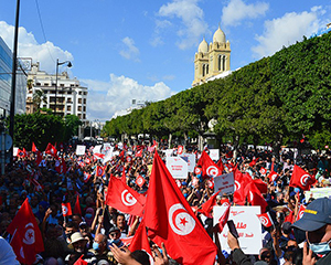 Many people protest in the streets while carrying signs and Tunisian flags.